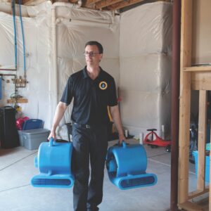 A water damage technician carrying two large blue industrial air movers into a basement to begin the drying process.