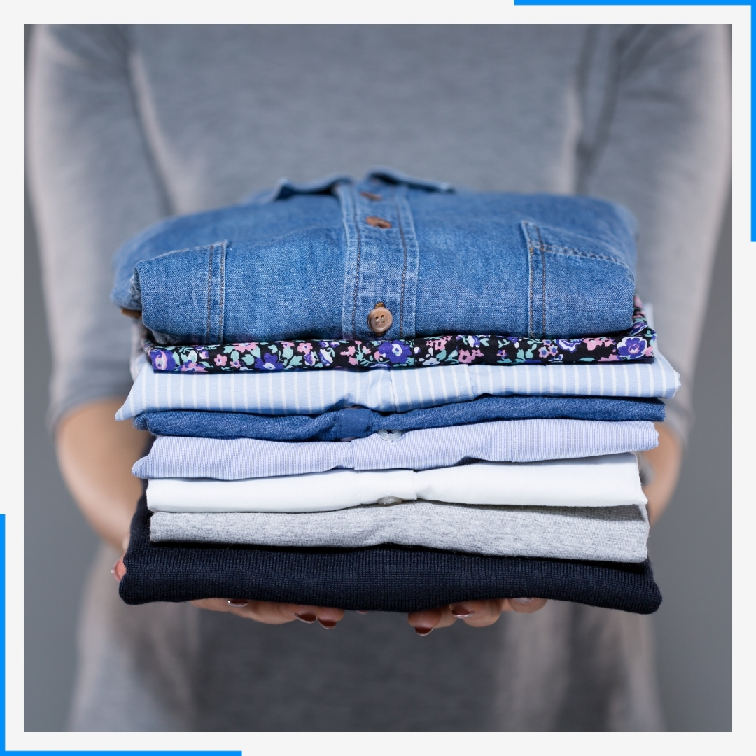 A person holds a neatly folded stack of clean clothes, including a denim shirt and several t-shirts.