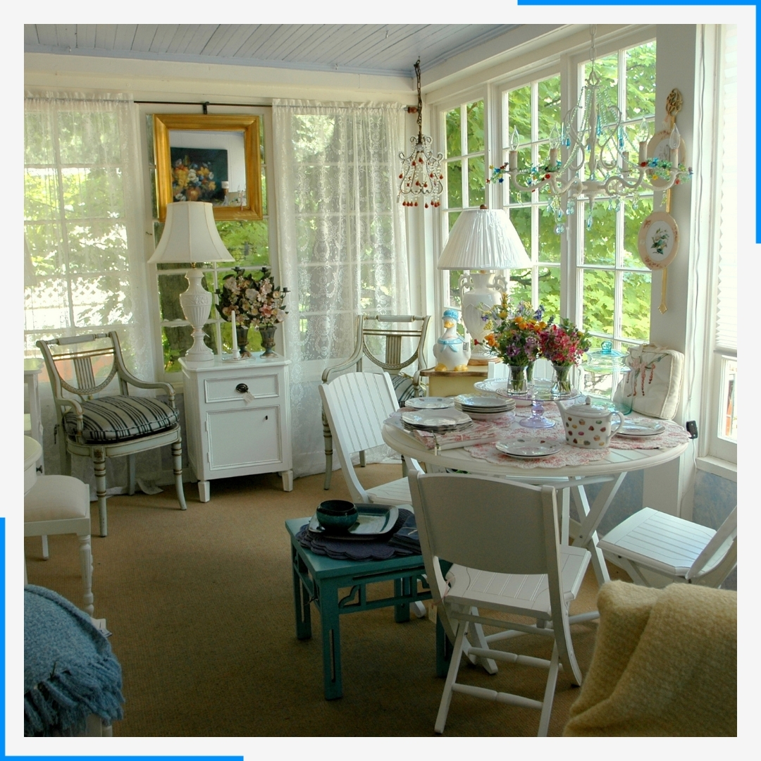 A brightly lit sunroom decorated in a shabby-chic style with white furniture and lace curtains.