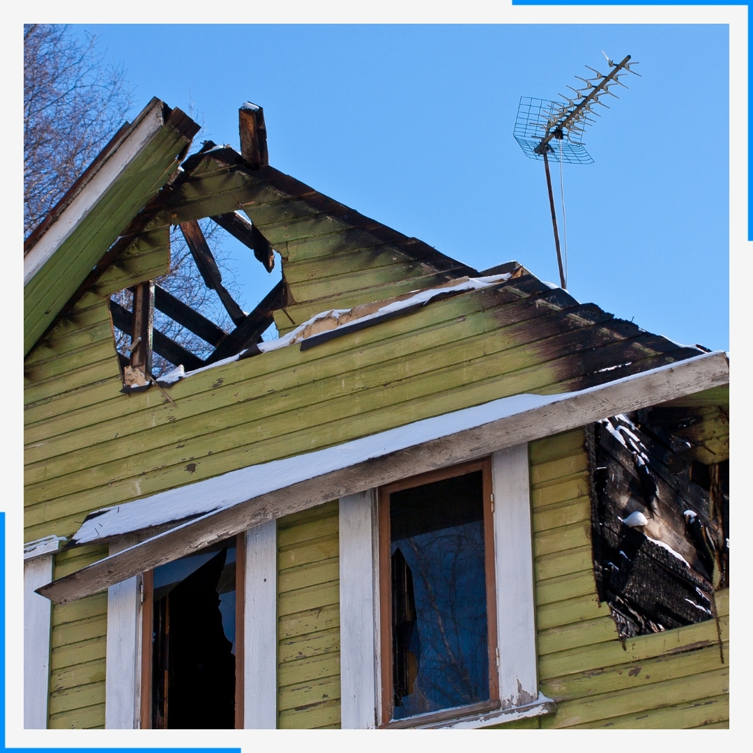 The upper story of a green house showing severe fire damage, with a burnt, collapsed roof and broken windows.