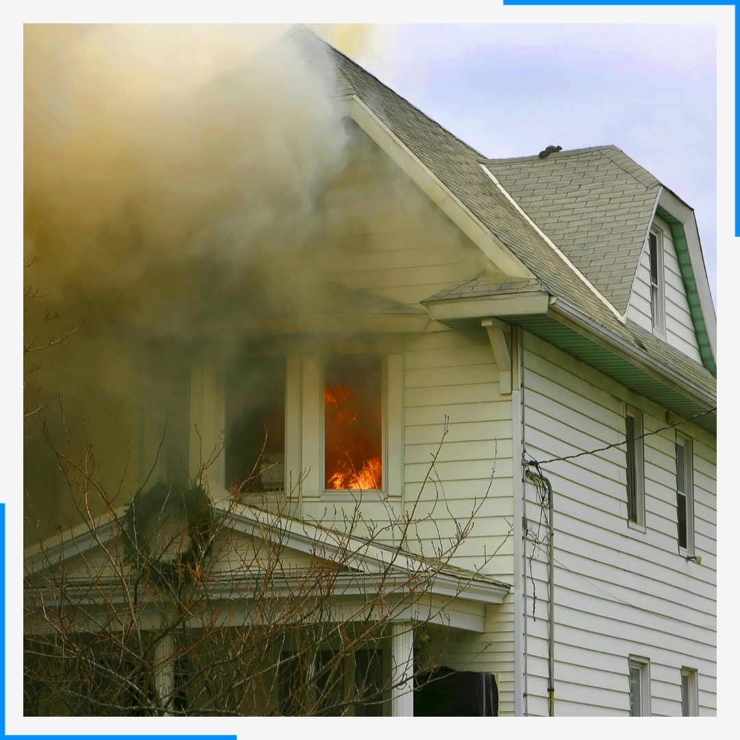 A white house with heavy smoke pouring out of the second story, and flames visible through a window.