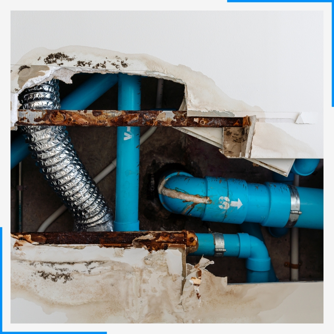 A hole in a water-damaged ceiling reveals rusty metal framing, blue PVC pipes, and a silver ventilation duct.