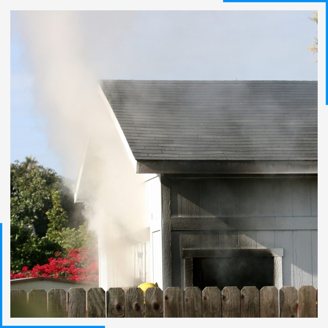 Smoke billows from a grey shed behind a wooden fence.