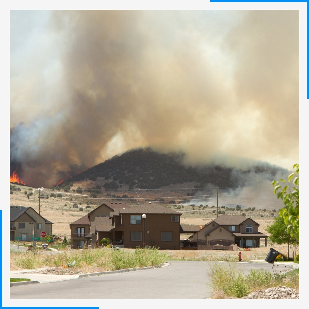 A suburban neighborhood sits at the base of a large hill where a wildfire is burning and creating a large smoke cloud.