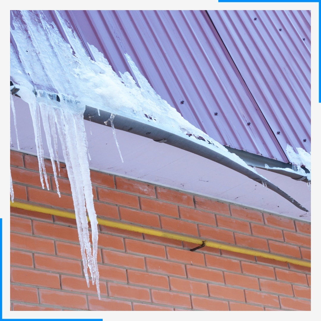Large icicles hang from the gutter of a building with a red metal roof, indicating an ice dam.